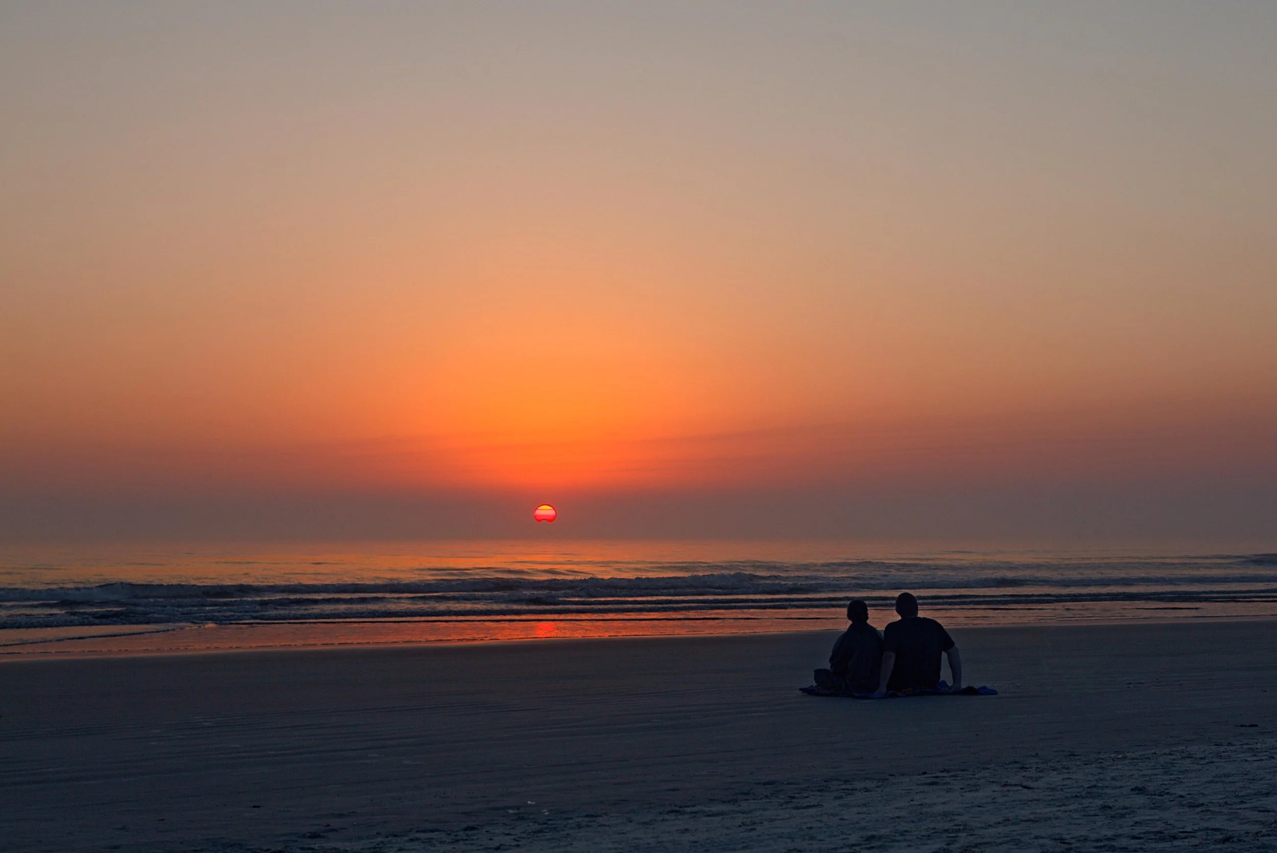 Two people sitting on Daytona Beach at sunrise, watching the sun over the ocean.