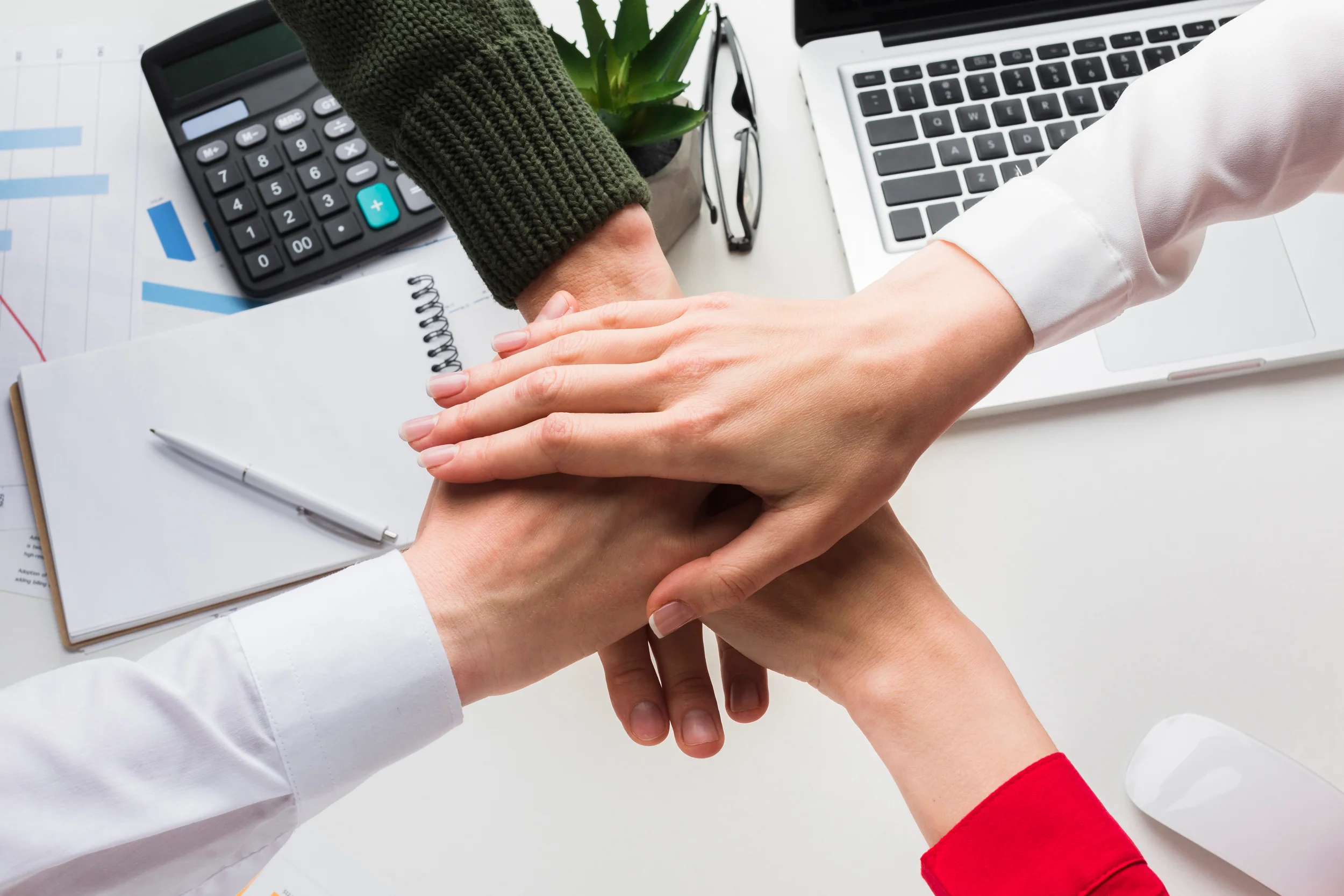 Stacked hands over a desk with a calculator and laptop