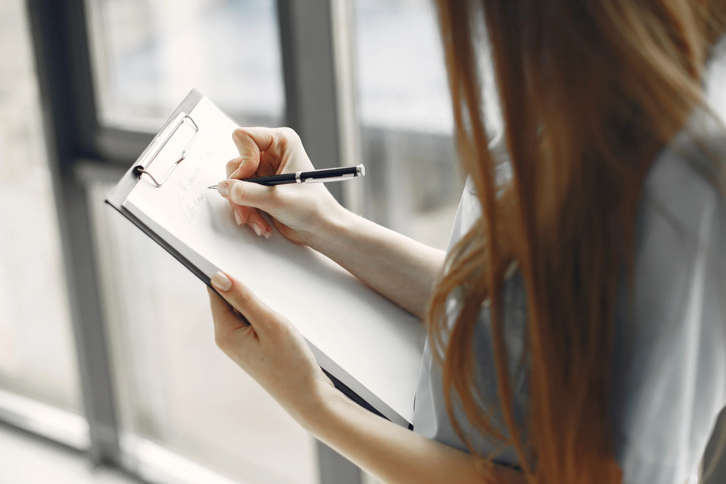 Woman writing on a clipboard by a window