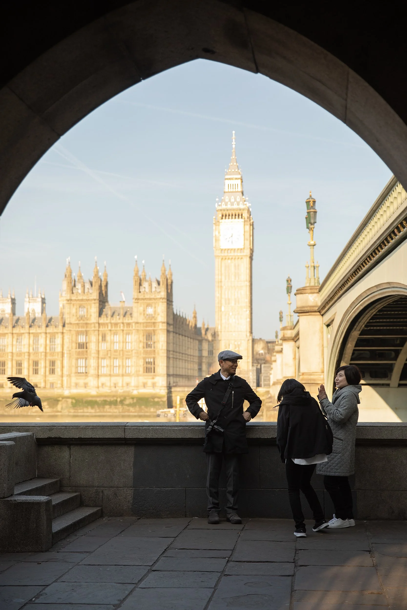 a family of three against Big Ben seen through an arch from the other side of River Thames