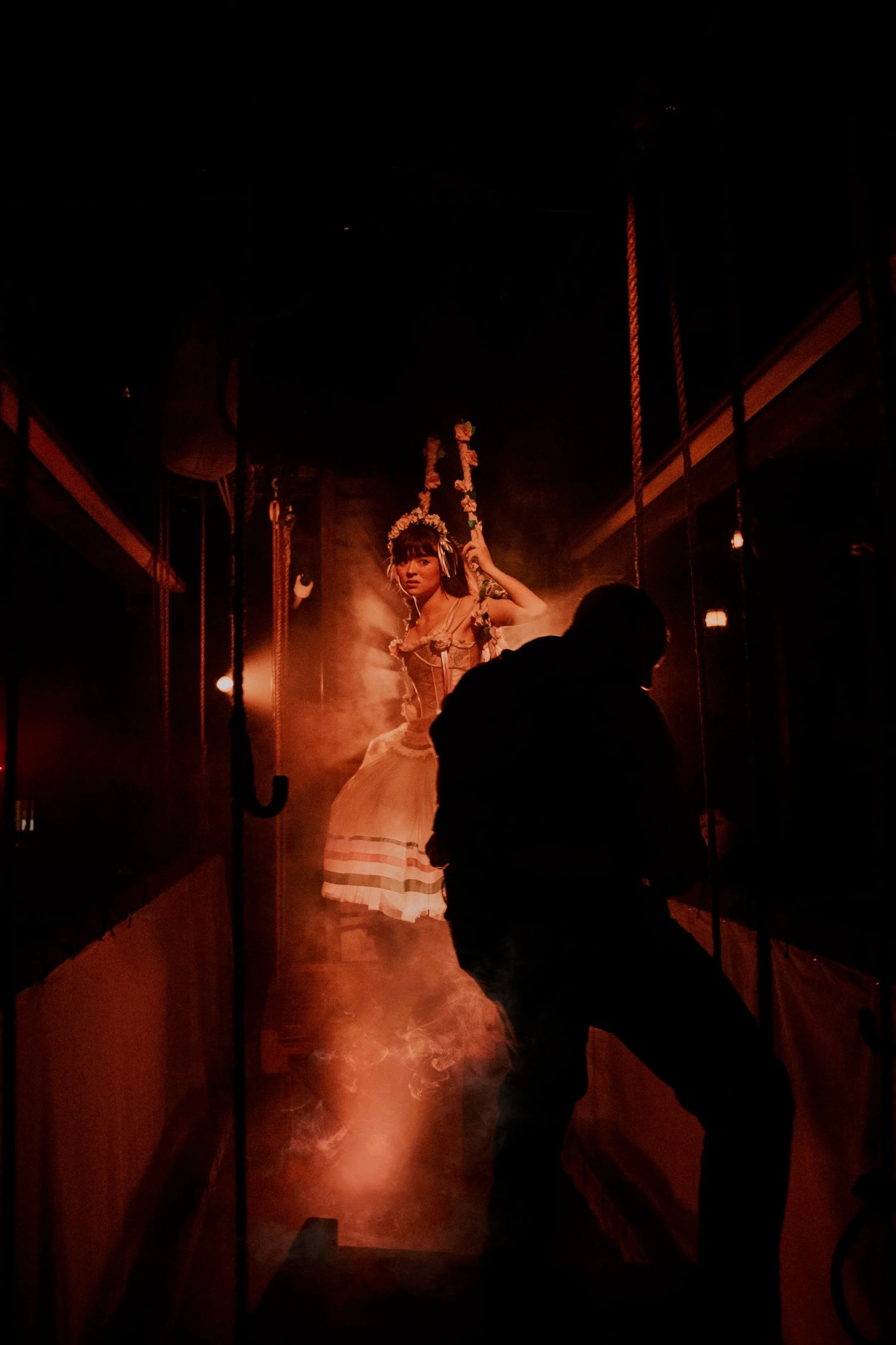 A woman in a dress sits in the rafters of a theatre on a swing.