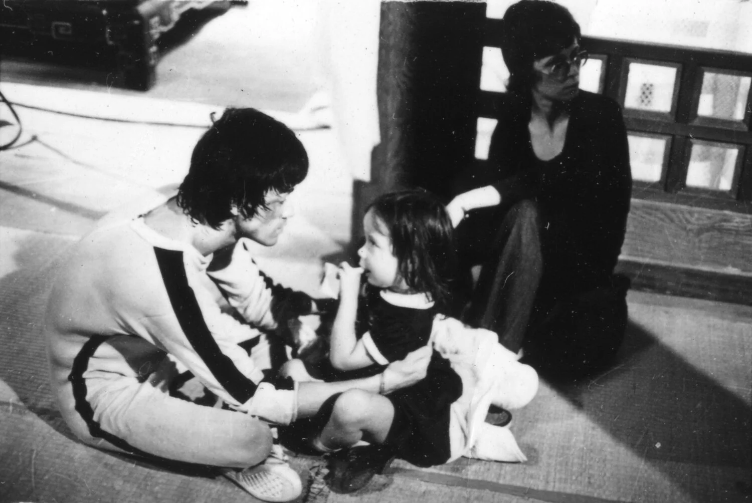 A black and white photograph of a mixed Asian man sitting cross-legged on the floor with his daughter. A white woman sits nearby. The man is wearing a jumpsuit with black stripes going down the sides