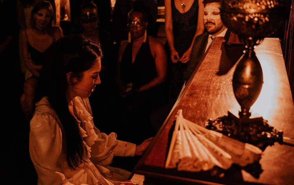 A woman plays the piano. Audience members watch wearing masks.