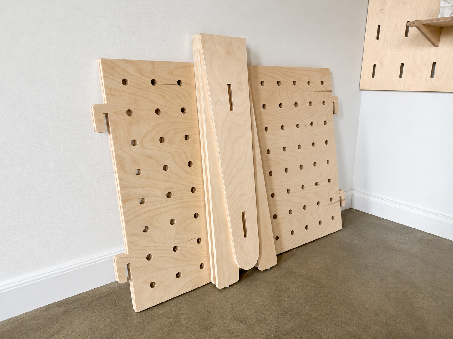 Flat-packed freestanding birch plywood pegboard display components leaning against a white wall before assembly