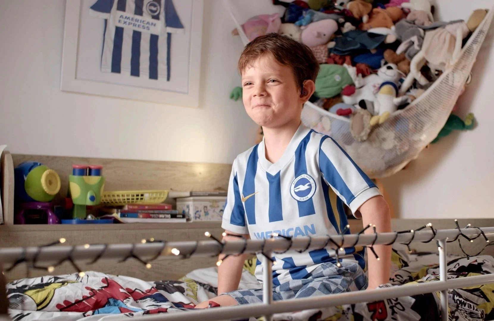 A young boy wearing a Brighton & Hove Albion football jersey sitting on a bed, smiling with a playful expression. The bed has colorful superhero-themed bedding. Behind him is a wall-mounted basket filled with plush toys, and a framed Brighton & Hove Albion football jersey is hanging on the wall.