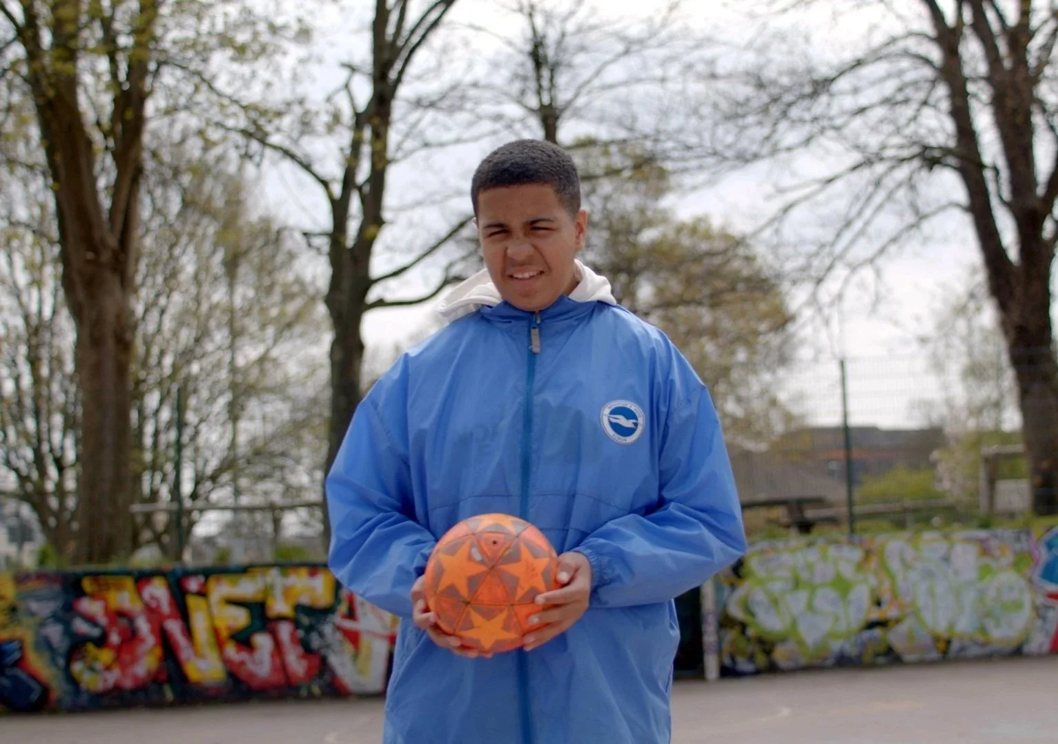 A young boy in a blue jacket holding an orange soccer ball with star patterns on an outdoor basketball court with colorful graffiti walls and leafless trees in the background.