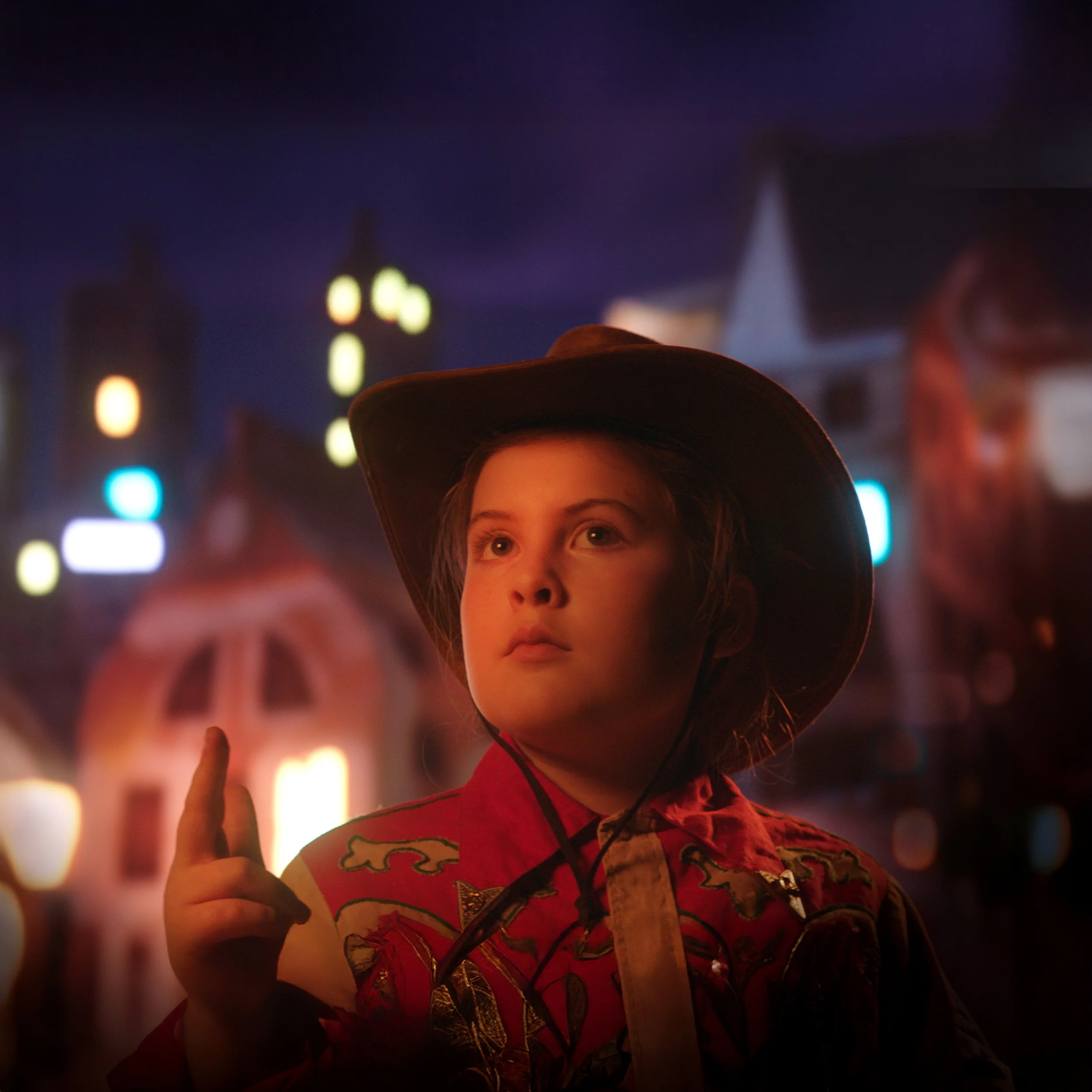 A young boy wearing a cowboy hat and a red jacket with a cartoon dragon pattern, standing at night in front of colorful blurred city lights.