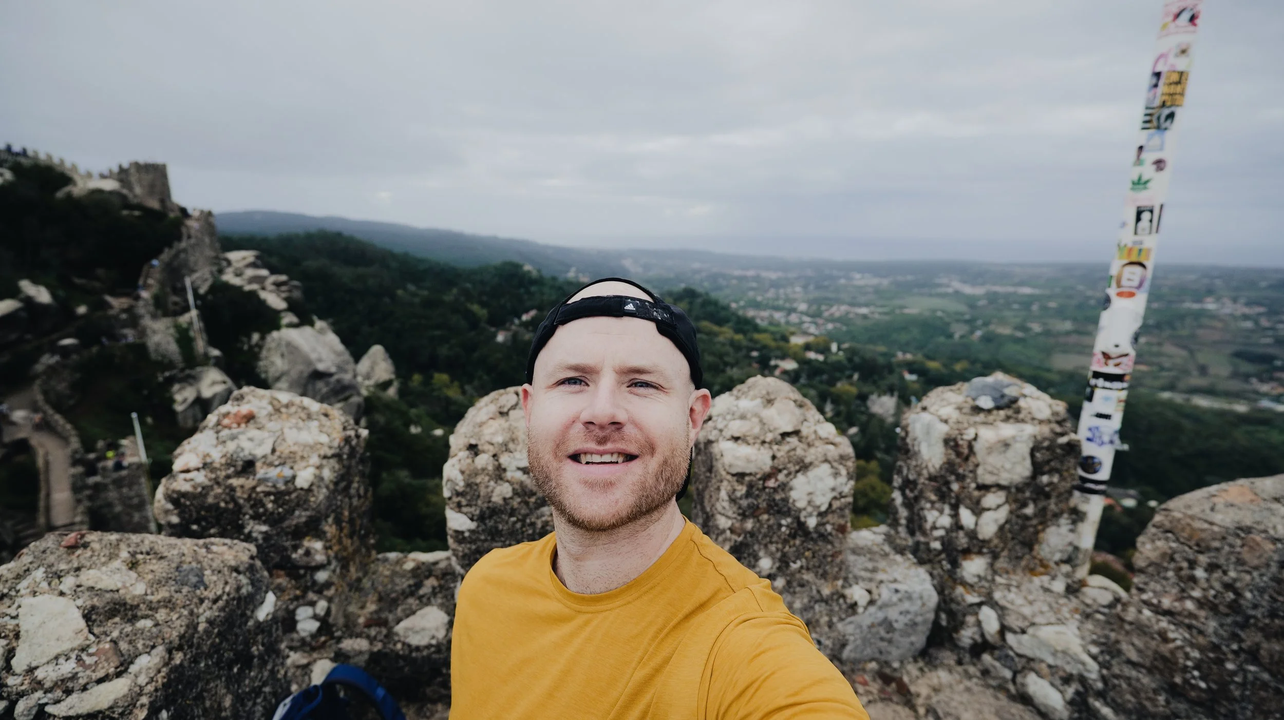 Man in a mustard yellow shirt and black cap taking a selfie on a stone viewpoint with scenic landscape in the background.