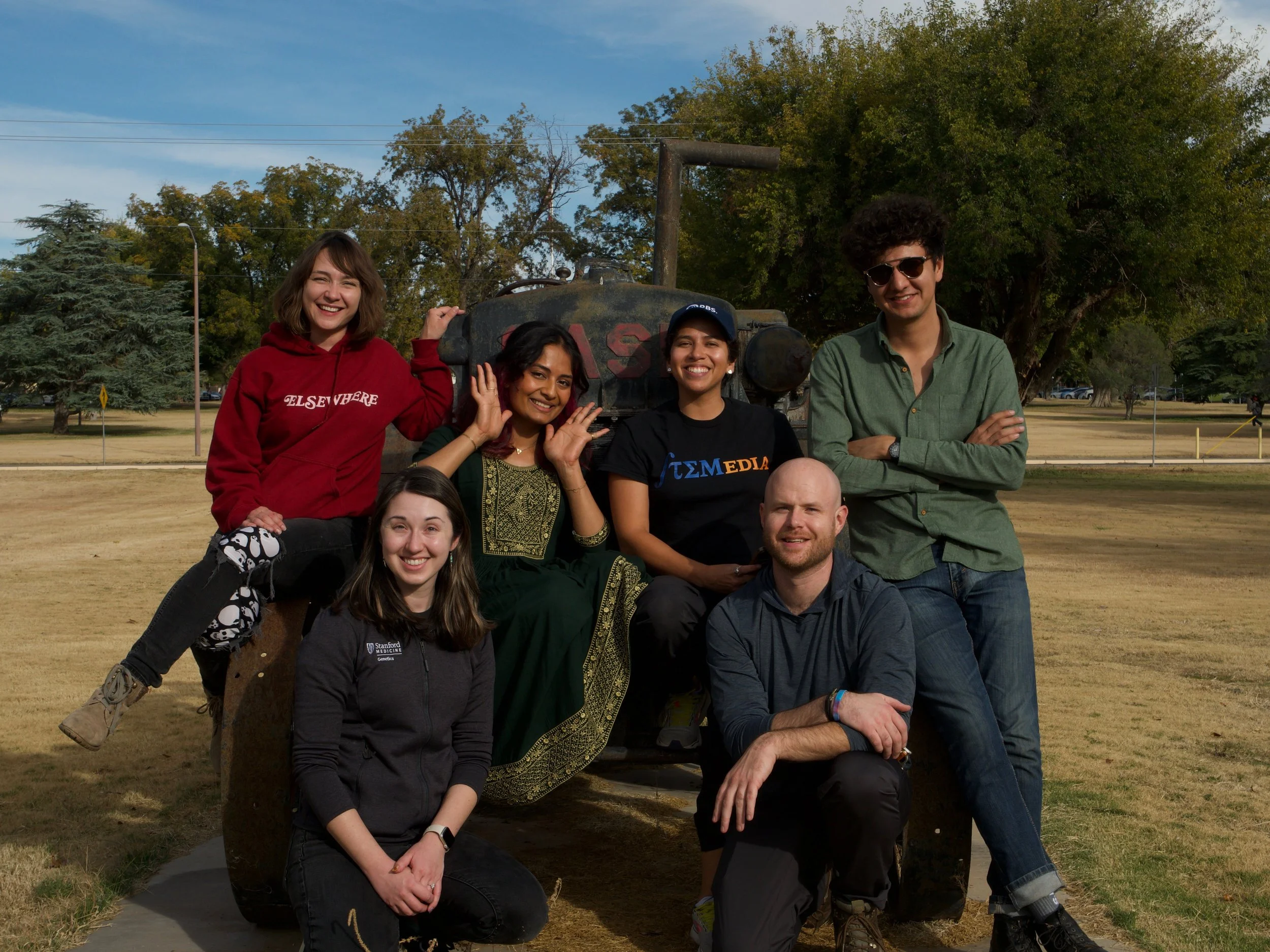 A group of six people smiling and posing outdoors in a park with trees in the background. They are sitting and standing around a large sculpture. The individuals are wearing casual clothing, and one person is wearing a traditional outfit.
