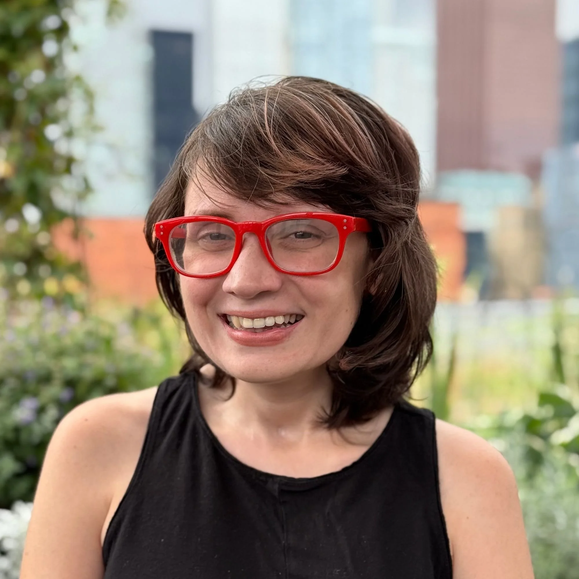 Keat headshot, smiling. Black shirt and red glasses. outside with blurry background
