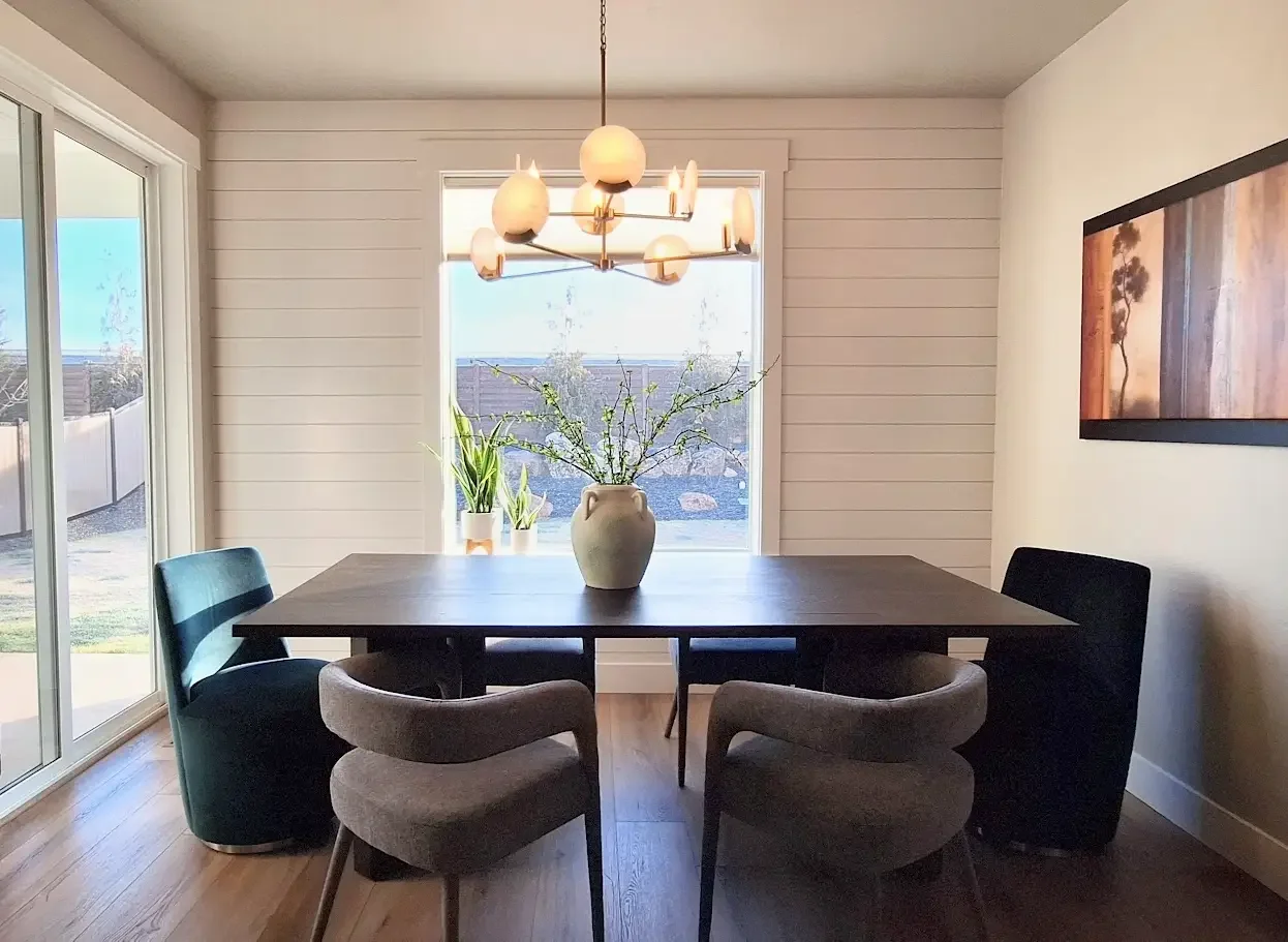 Dining room with a dark wood table, gray and black chairs, a beige vase with branches, two potted plants, and a chandelier with round lights, illuminated by sunlight through large windows.