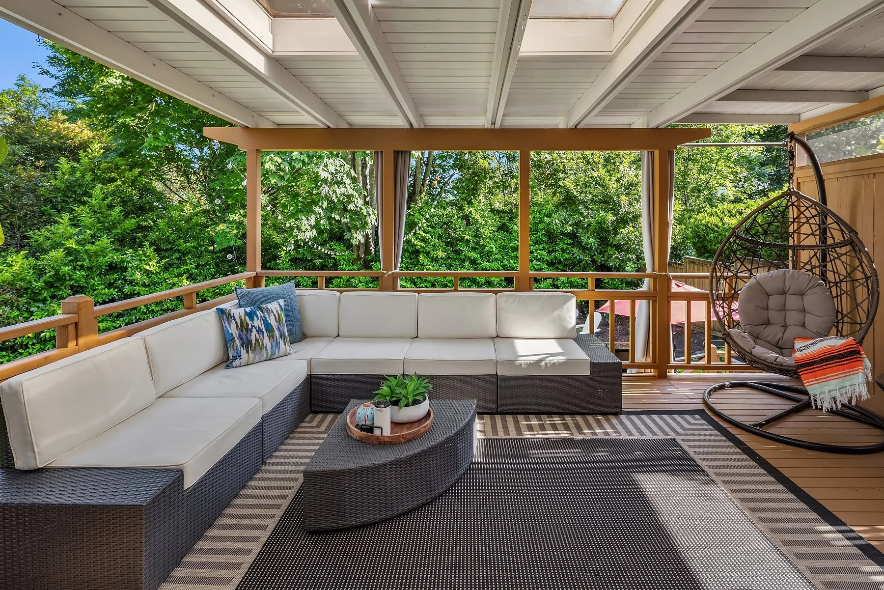 Covered outdoor porch with skylights, white cushioned sectional sofa, and patio egg swing chair overlooking a private lush green garden.