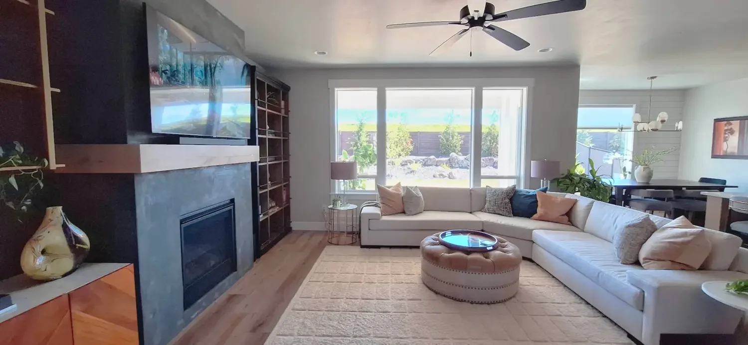 Living room with large window, beige sectional sofa with multiple pillows, round tufted ottoman with tray, two side tables with lamps, black fireplace, wall-mounted TV, built-in bookshelf, and various indoor plants; dining area in the background with a black table and chairs, and decorative lighting fixtures.
