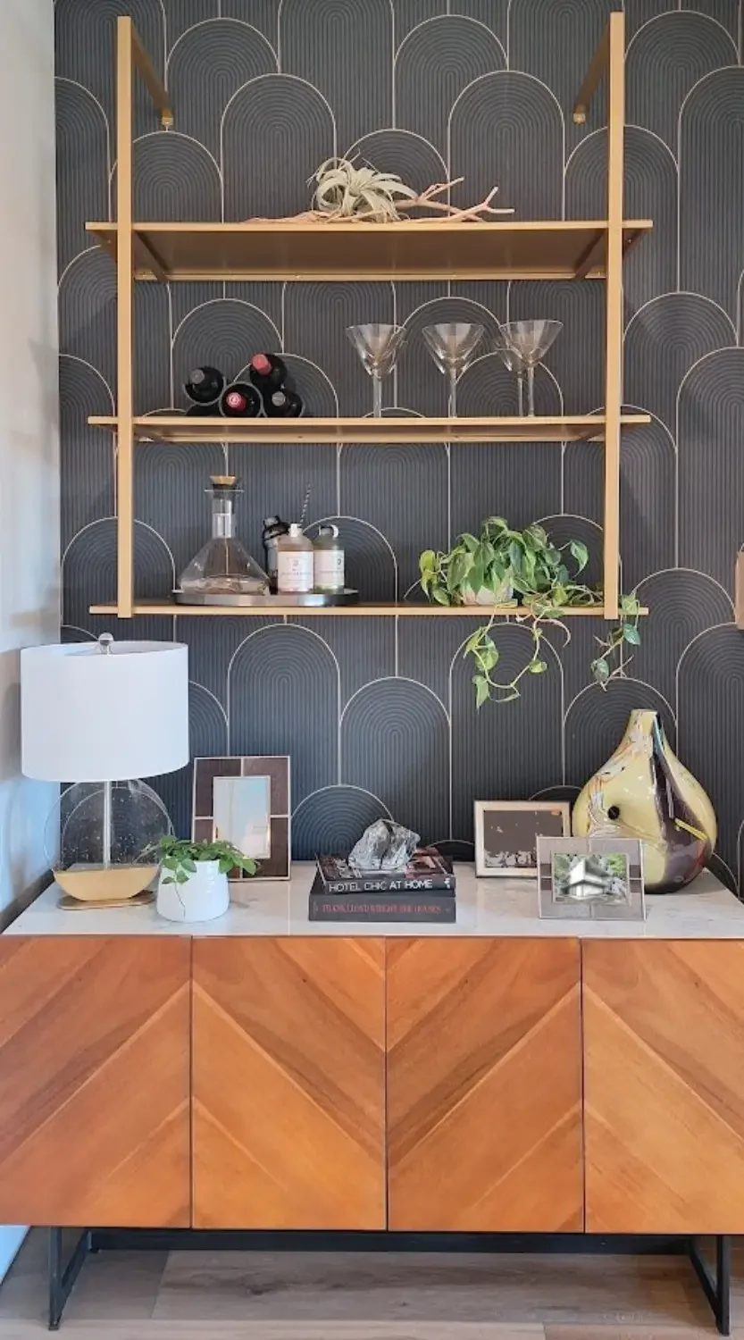 Interior decor shelf with wine bottles, glasses, plants, and framed photos on a wooden console against a dark patterned wall.
