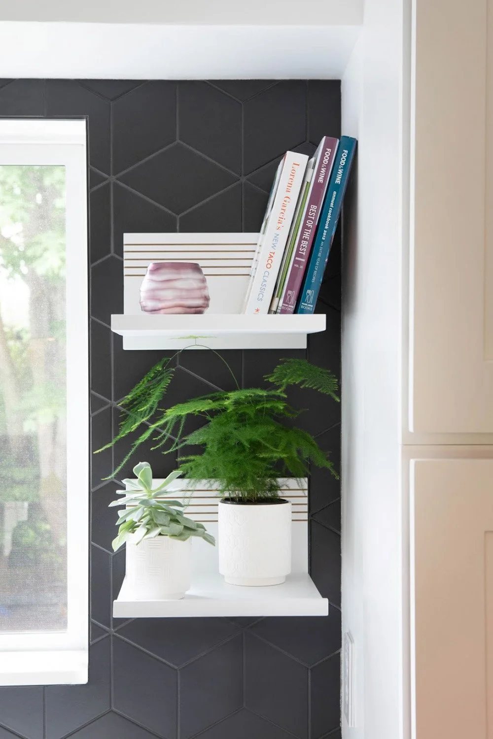 Wall‑mounted custom shelves with cookbooks and asparagus fern against black rhombus tile wall beside large window.