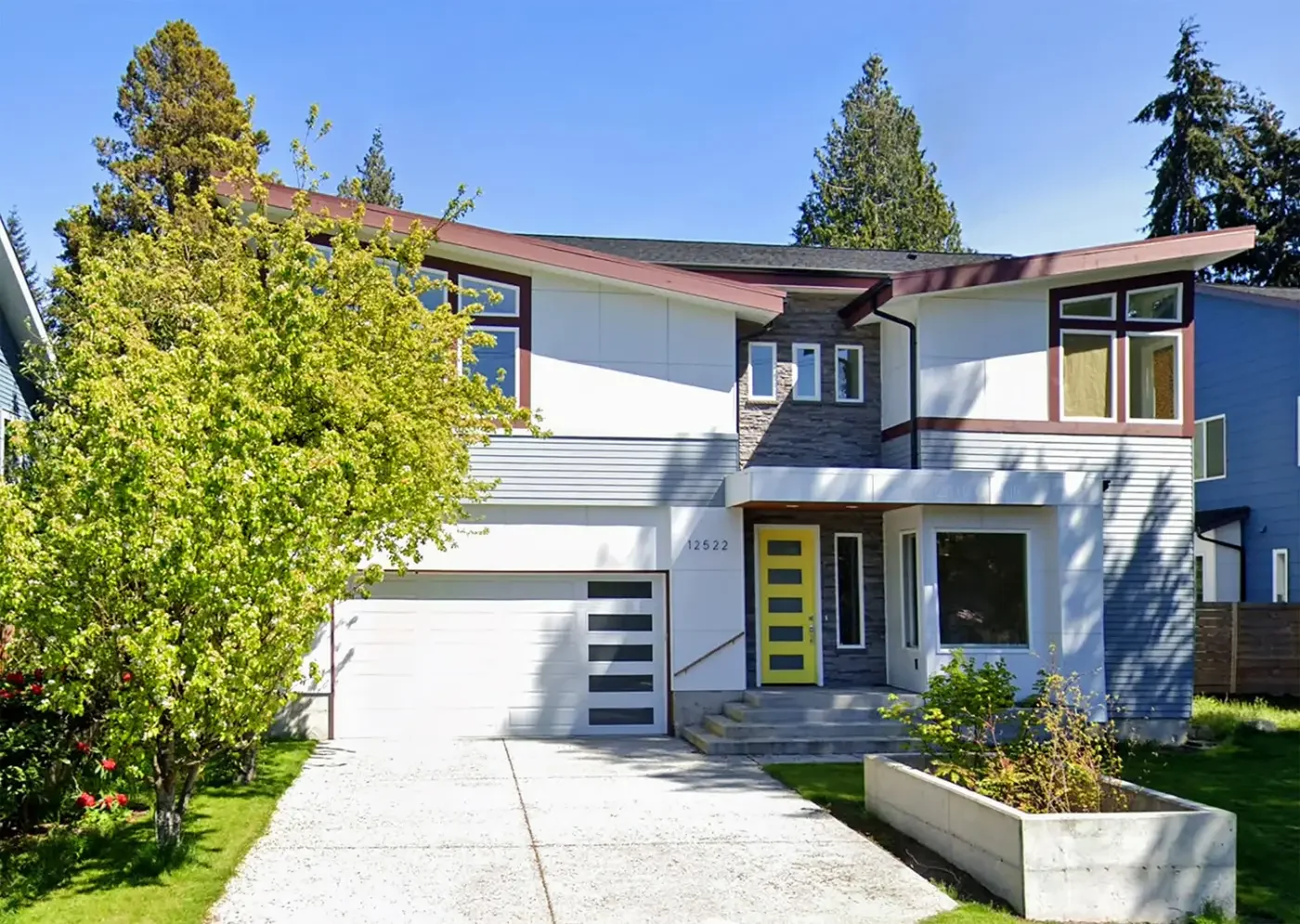 Modern two-story house with white and gray exterior, yellow front door, large windows, driveway, and surrounding greenery under a clear blue sky.