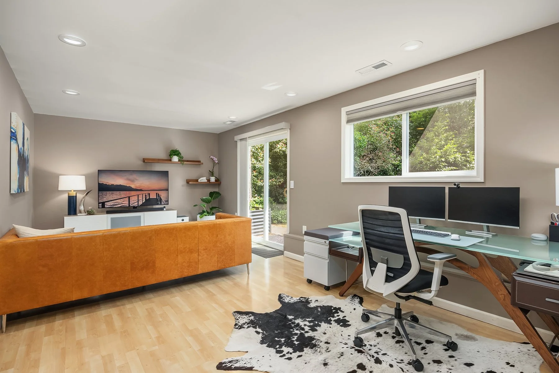 Modern flex room for office and family use with mahogany wood and glass desk, Herman Miller Aeron chair, cowhide rug, and CB2 cognac leather sofa.