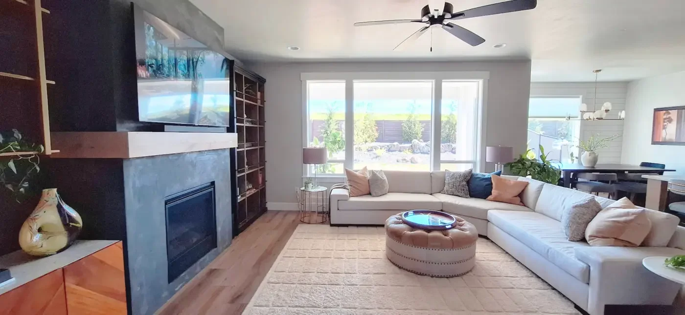 Living room with large window, beige sectional sofa with multiple pillows, round tufted ottoman with tray, two side tables with lamps, black fireplace, wall-mounted TV, built-in bookshelf, and various indoor plants; dining area in the background with a black table and chairs, and decorative lighting fixtures.