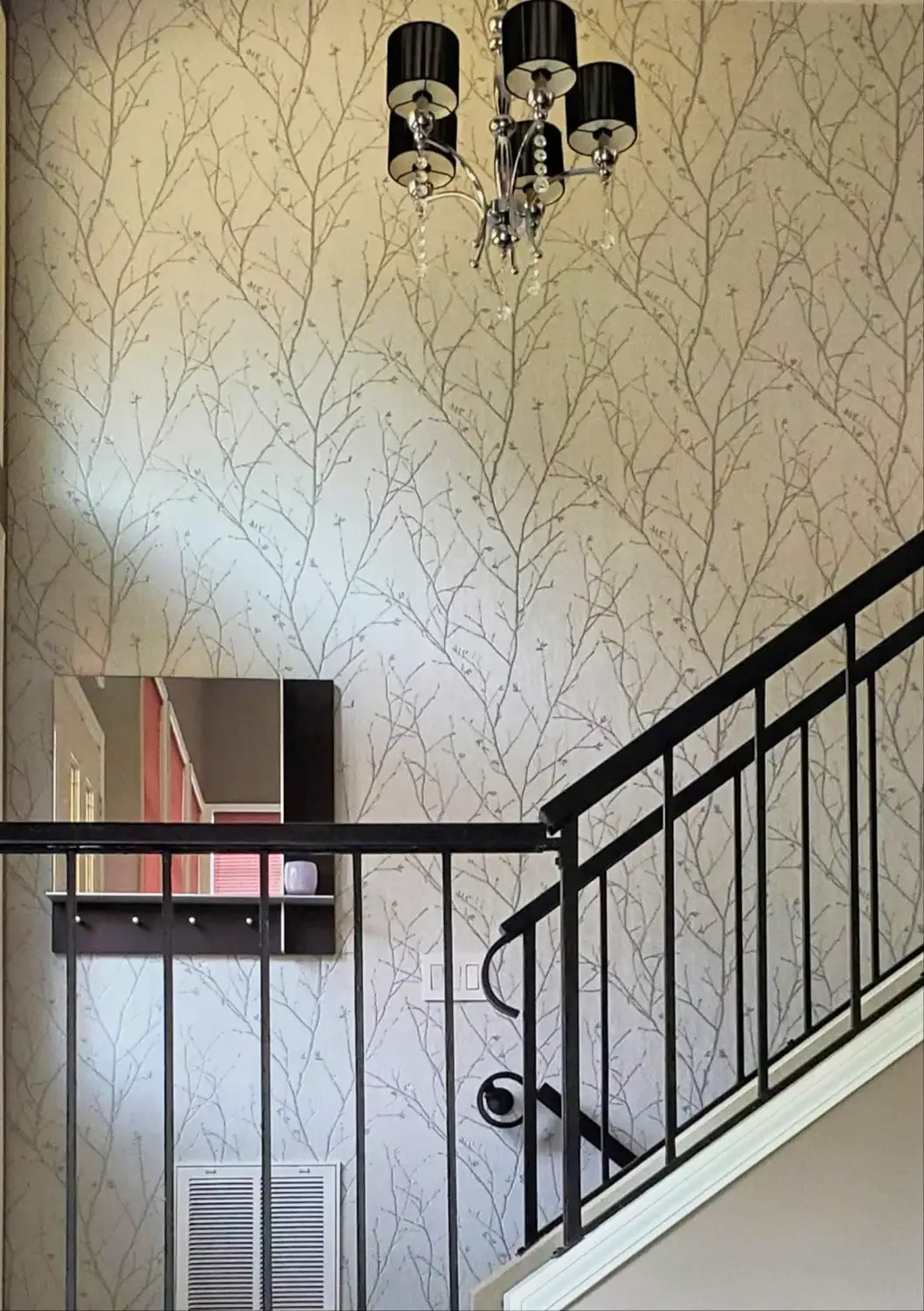 Staircase landing with Graham & Brown Water Silk Sprig Silver wallpaper, black iron railing, mirror, shelf, and chandelier—modern interior design in Meridian home.