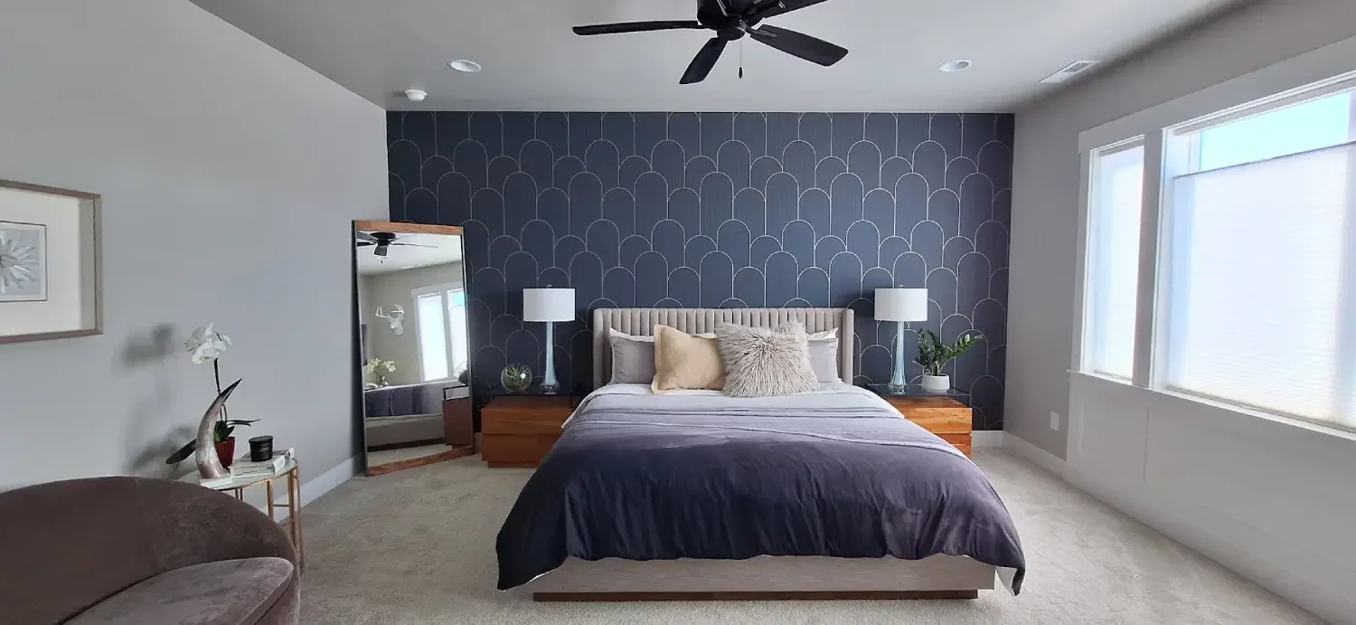A modern bedroom with a navy blue accent wall featuring gold geometric patterns. The bed has a beige upholstered headboard, and is adorned with pillows and a gray blanket. Two white bedside lamps sit on wooden nightstands on either side of the bed. To the left, there is a full-length mirror and a window with white blinds on the right, letting in natural light.