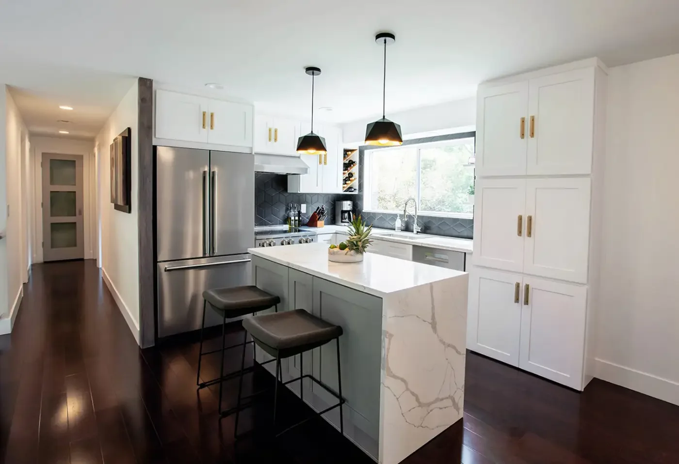 Modern kitchen with white cabinets, a central white marble island, black pendant lights, stainless steel appliances, and a large window overlooking trees.