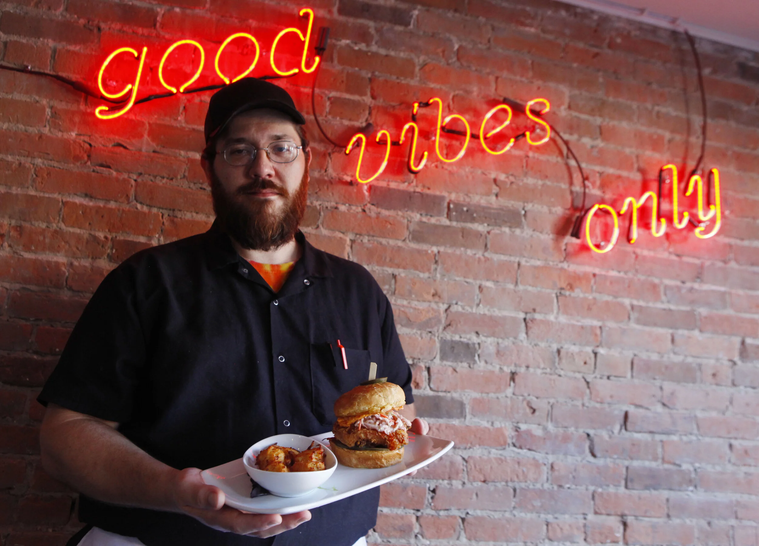 James Cannon, head chef at One13 North restaurant in Crown Point, poses for a portrait on July 23, 2019 underneath an electric sign reading ‘Good Vibes Only.’ Cannon is pictured with a fried chicken sandwich with a side of tater tots.