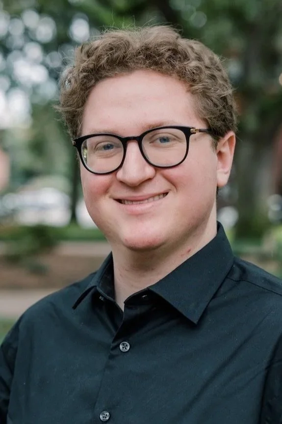 A young man with curly light brown hair, wearing glasses and a black button-up shirt, standing outdoors in a park with blurred trees in the background.
