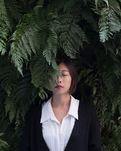 Ferns drape in front of a woman's face as she stands, eyes closed.