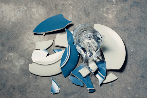 A blue ceramic plate has smashed on a cement floor. A glass of water is falling to join it.