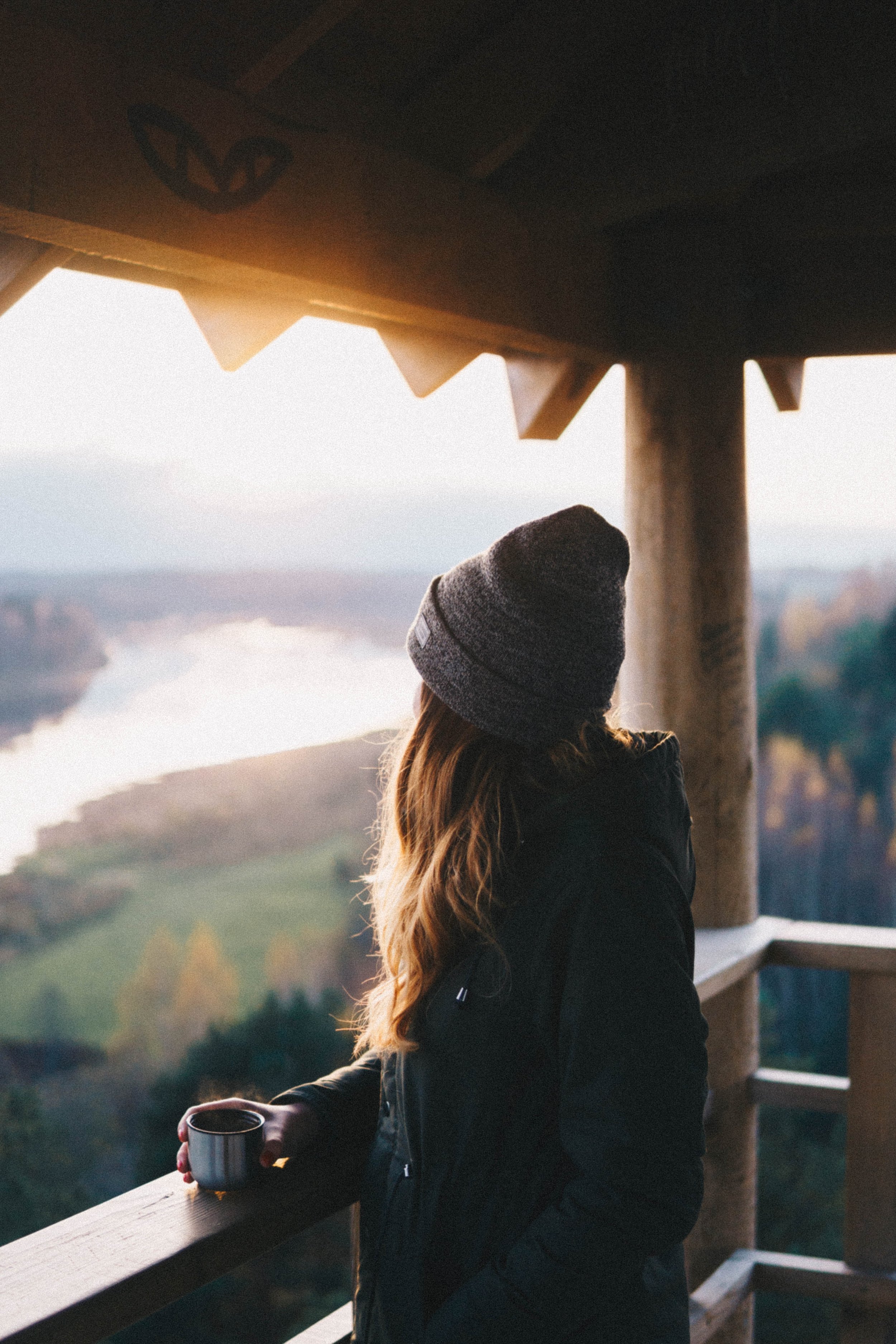 A woman with a mug standing at a railing, gazing out at a mountain valley