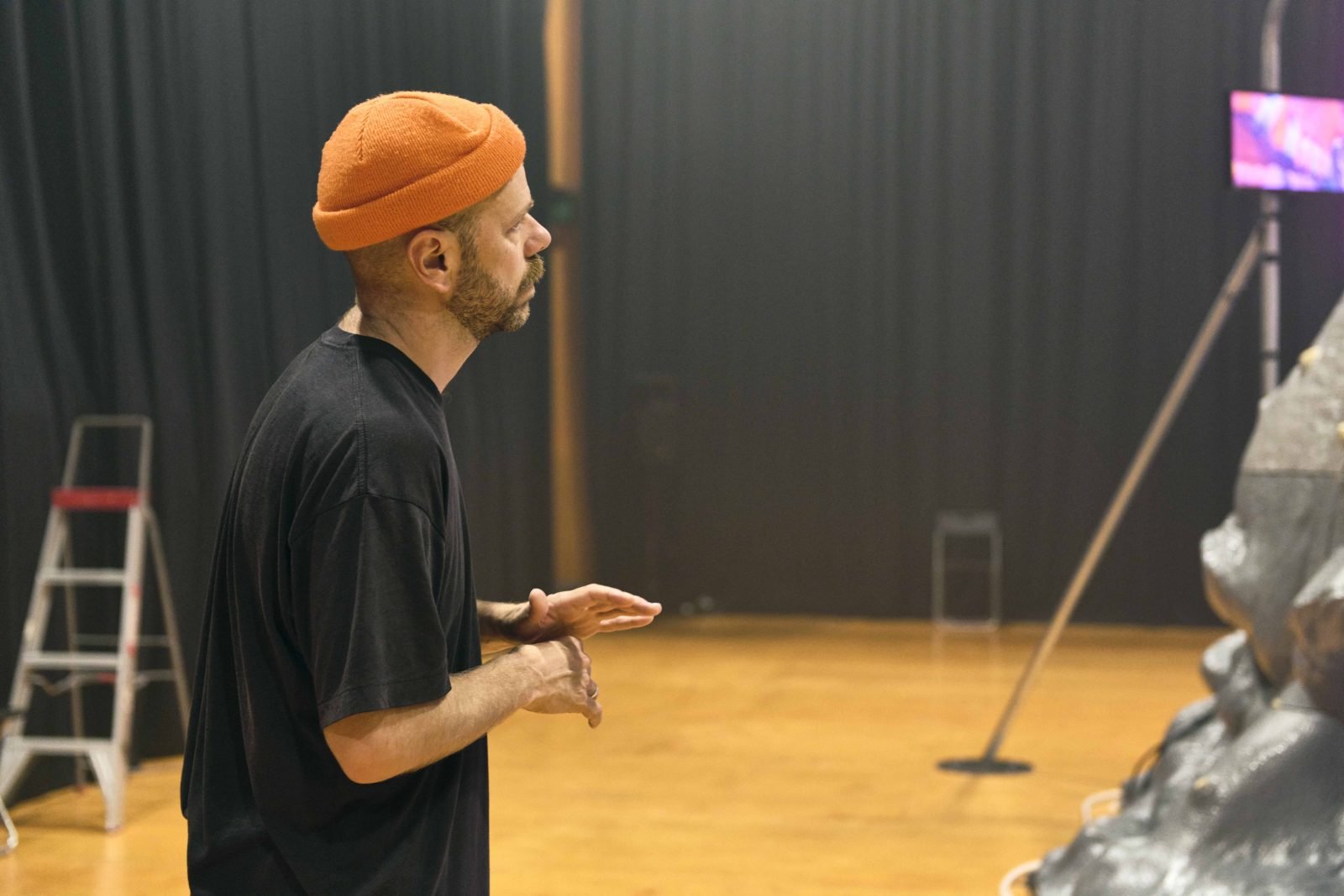 Antony Hamilton, stands in a rehearsal room, looking off to the side. Looking at a lard set piece out of frame.