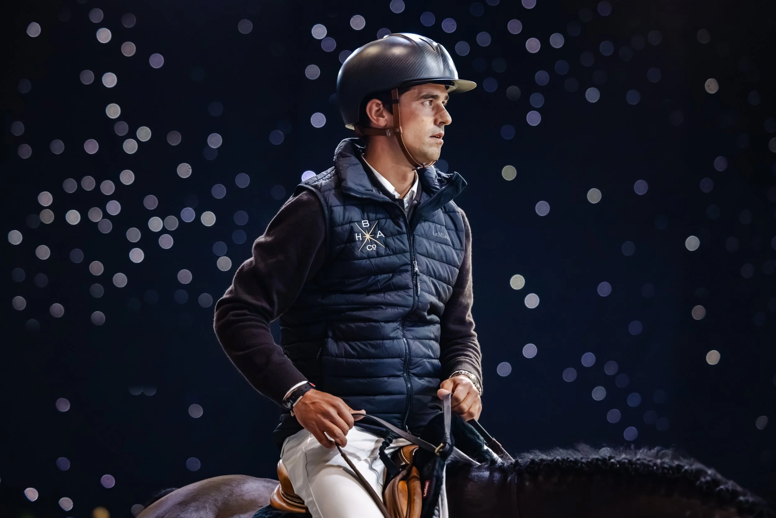 A man wearing a black riding helmet and a navy puffer vest sits on a horse at night, with a background of blurred circular lights.