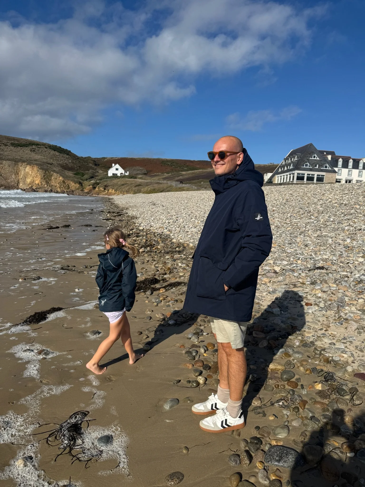 Sven Cichowicz mit Sonnenbrille an einem Strand, mit Küste und Gebäuden im Hintergrund.
