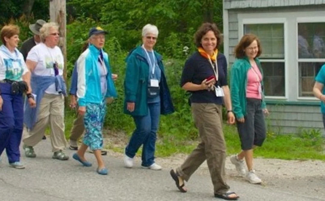 A group of eight women walking outdoors on a sidewalk next to a house, dressed casually with some wearing jackets and carrying backpacks or water bottles.