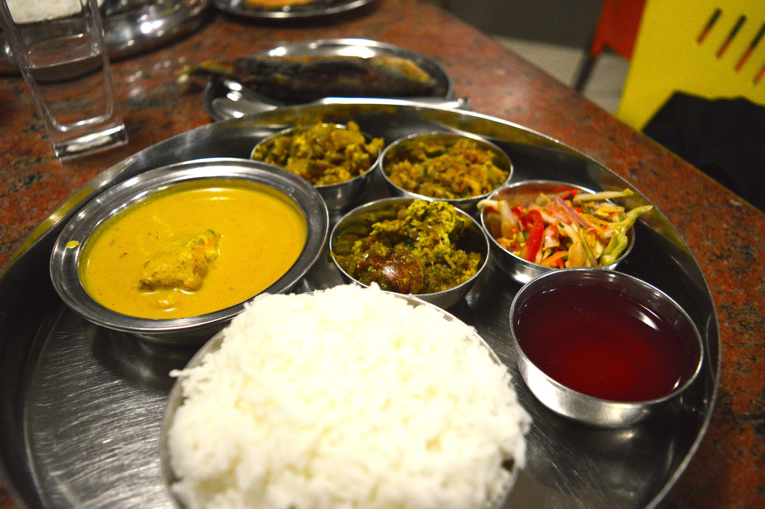  市場近くの食堂で食べた焼きサバ付きのタリー（定食）。左上から時計回りに、フィッシュカレー、ナス、アサリ（下）、桜えびのおかず、サラダ、消化を助けるコーカムカディ、ご飯。奥は焼きサバ。400円弱。 