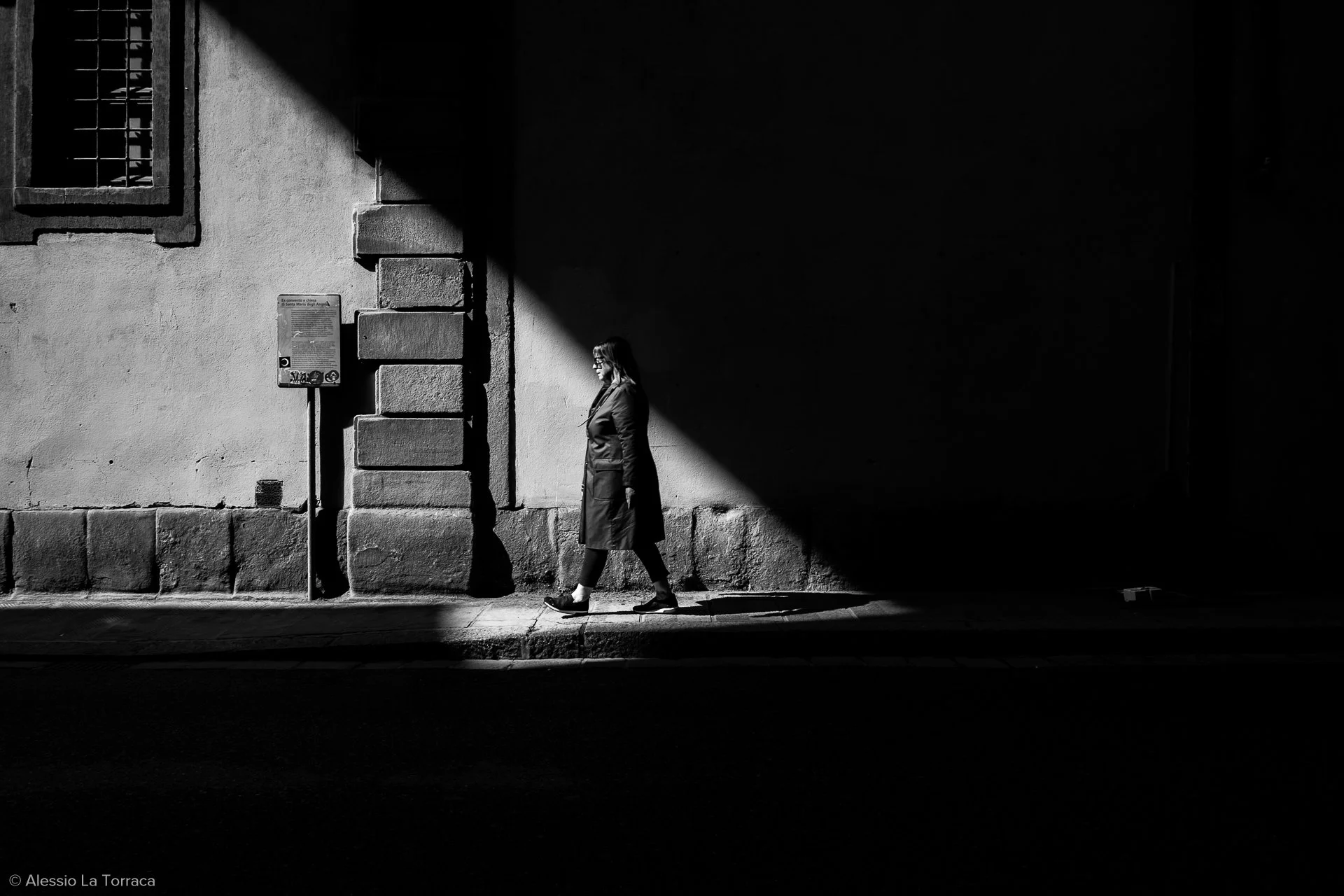 A woman walking on a city sidewalk in sunlight, with a large shadow covering part of the wall and ground, in black and white.