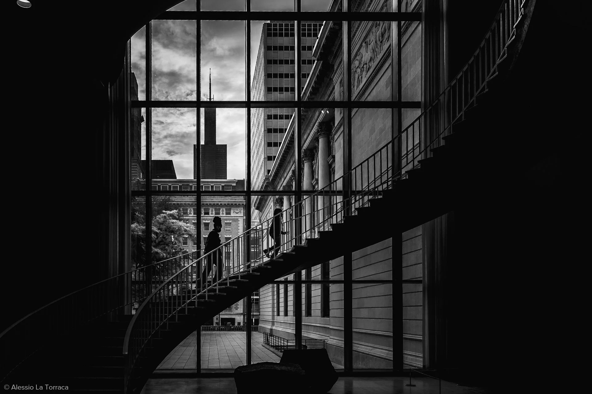 Silhouetted people walking upstairs inside a building with a large window showing city buildings and a cloudy sky outside.
