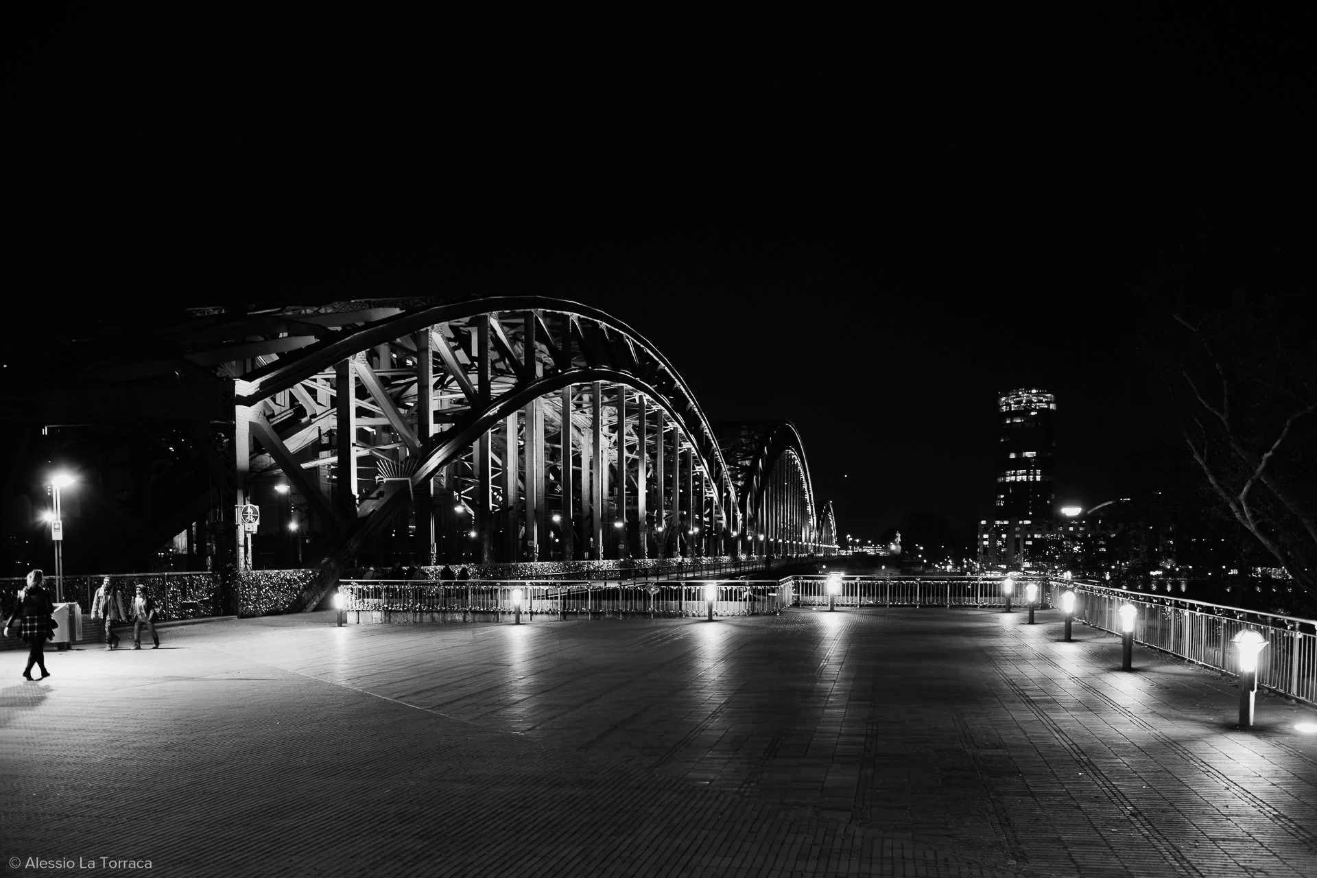 Nighttime view of a bridge with illuminated arches and city skyline in the background, captured in black and white.