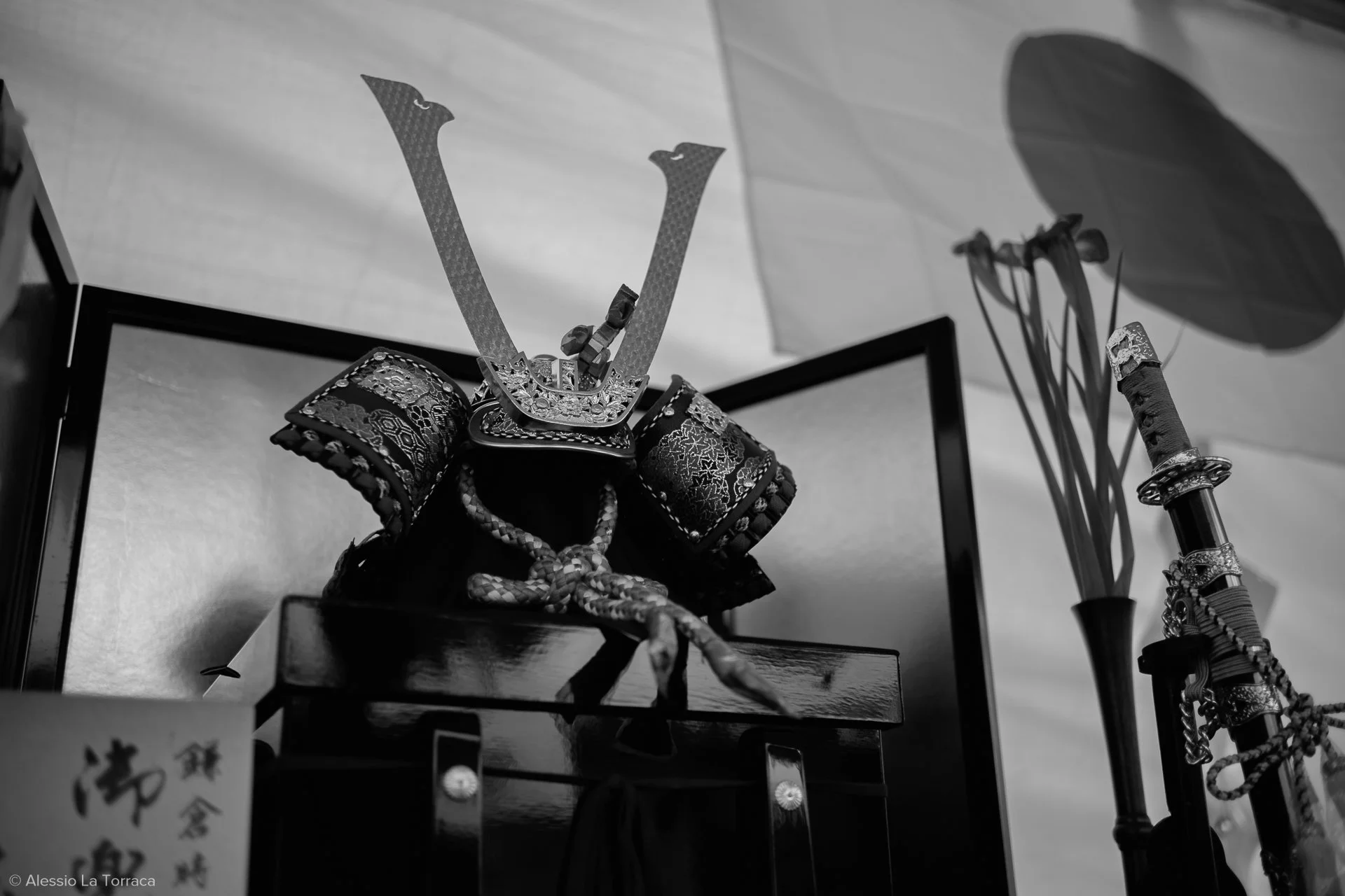 Black and white image of a miniature traditional Japanese samurai armor with a helmet, stand, and sword, displayed on a wooden surface with a parasol in the background.