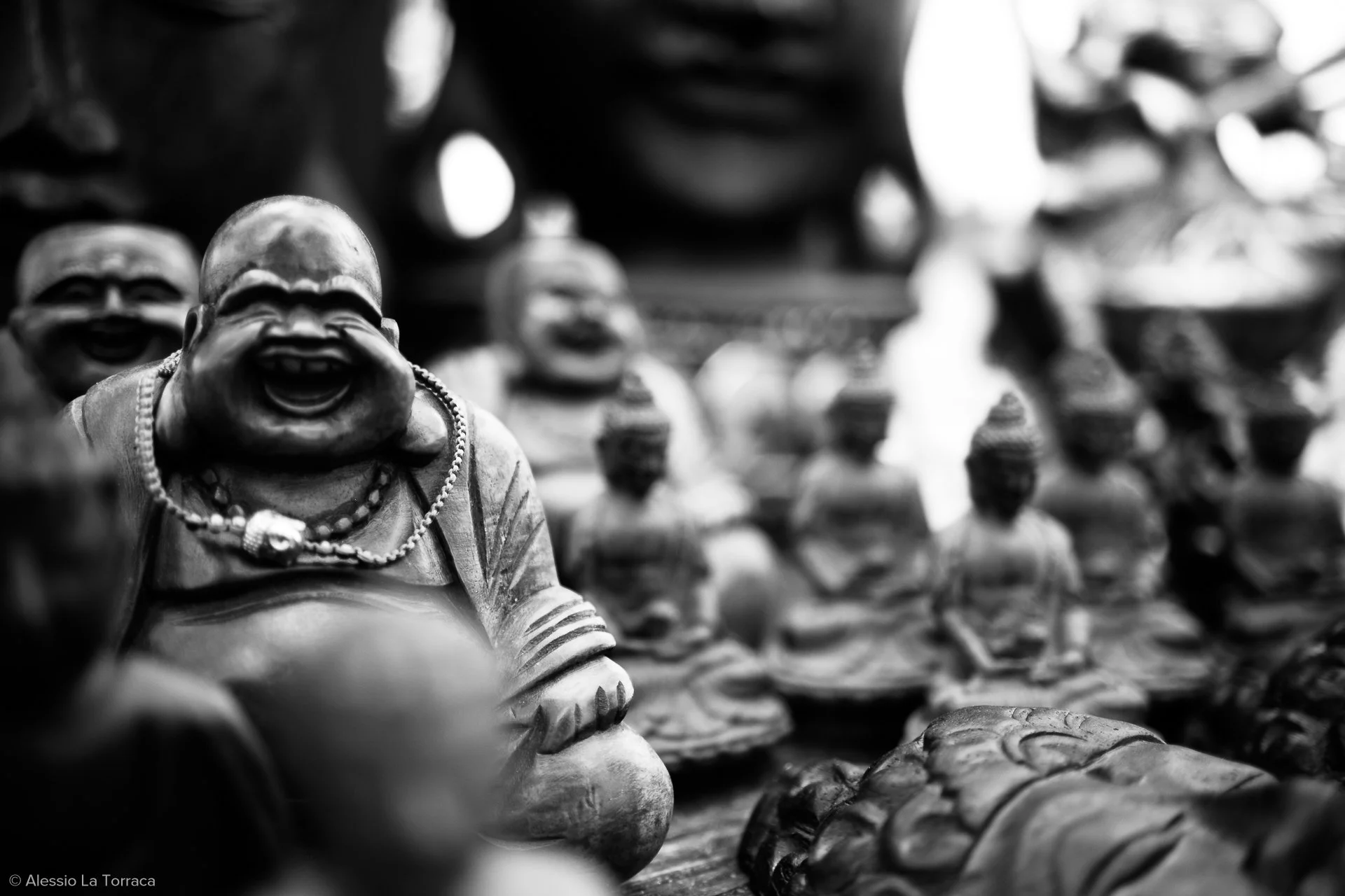 Black and white photo of multiple smiling Buddha statues, with a focus on a laughing Buddha with a necklace in the foreground and several others in the background.