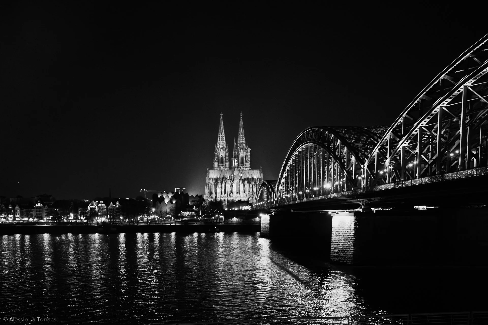 Night view of Cologne Cathedral and Hohenzollern Bridge over the Rhine River in Cologne, Germany, illuminated with city lights.