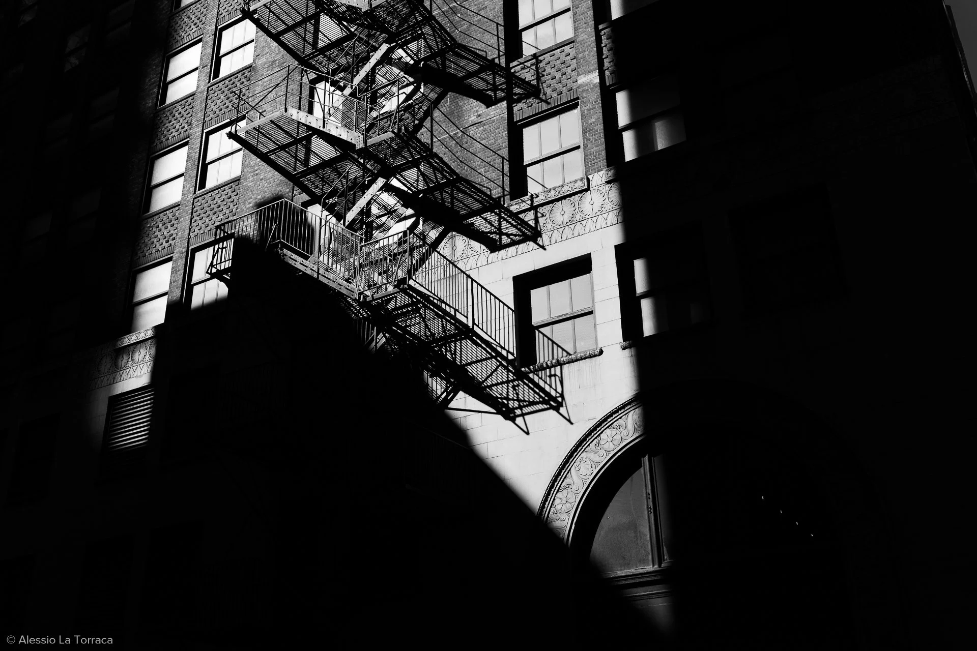Black and white photo of a fire escape on the side of a building, with light and shadow creating a contrast on the facade.