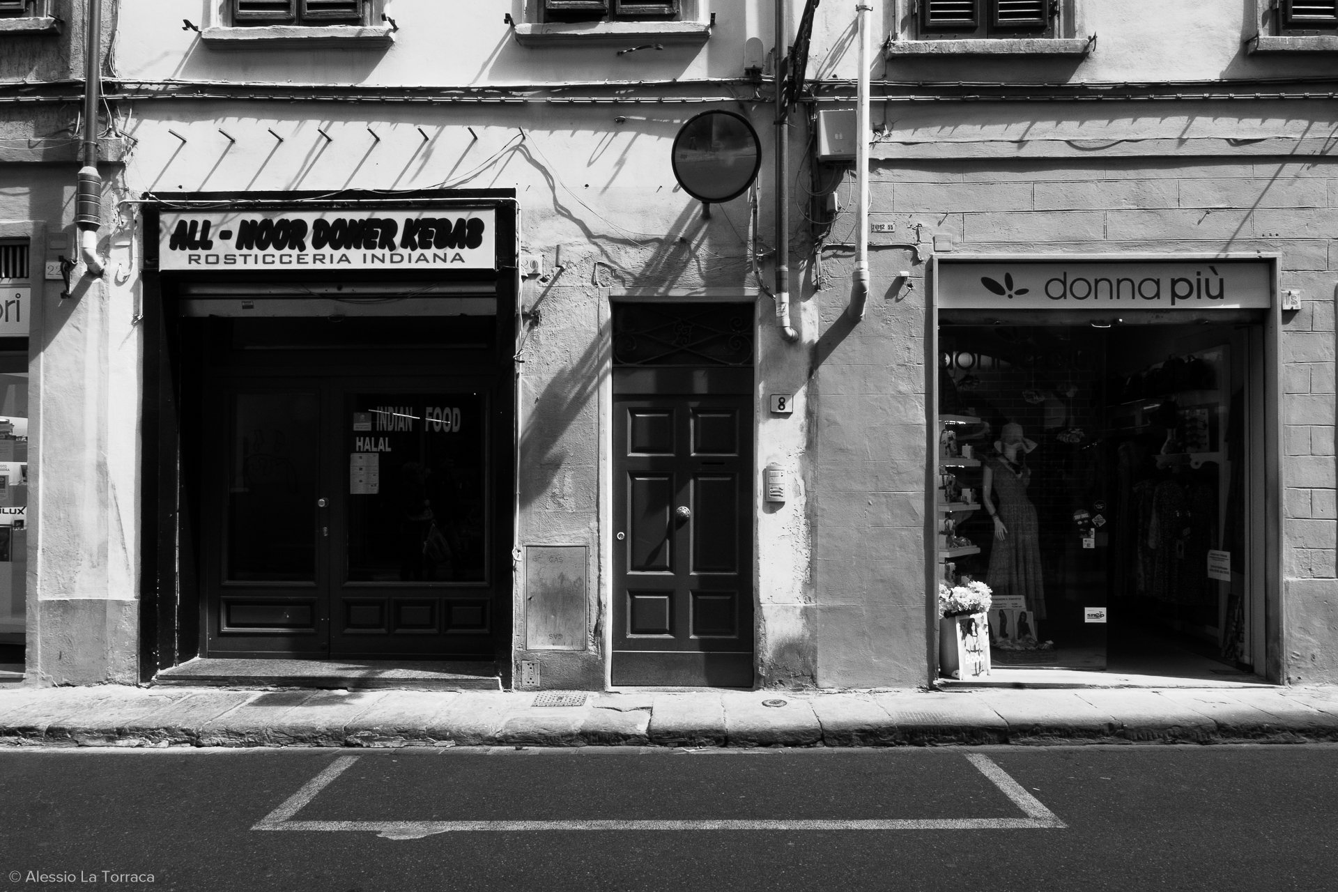 Black and white photo of a building facade with three doors and signs. The left door has a sign above it that reads 'ALL - NOOR DINER KEBAB ROSTICCERIA INDIANA.' The middle door is plain with a door handle. The right door is a shop named 'donna piu,' with displays of clothing visible inside.