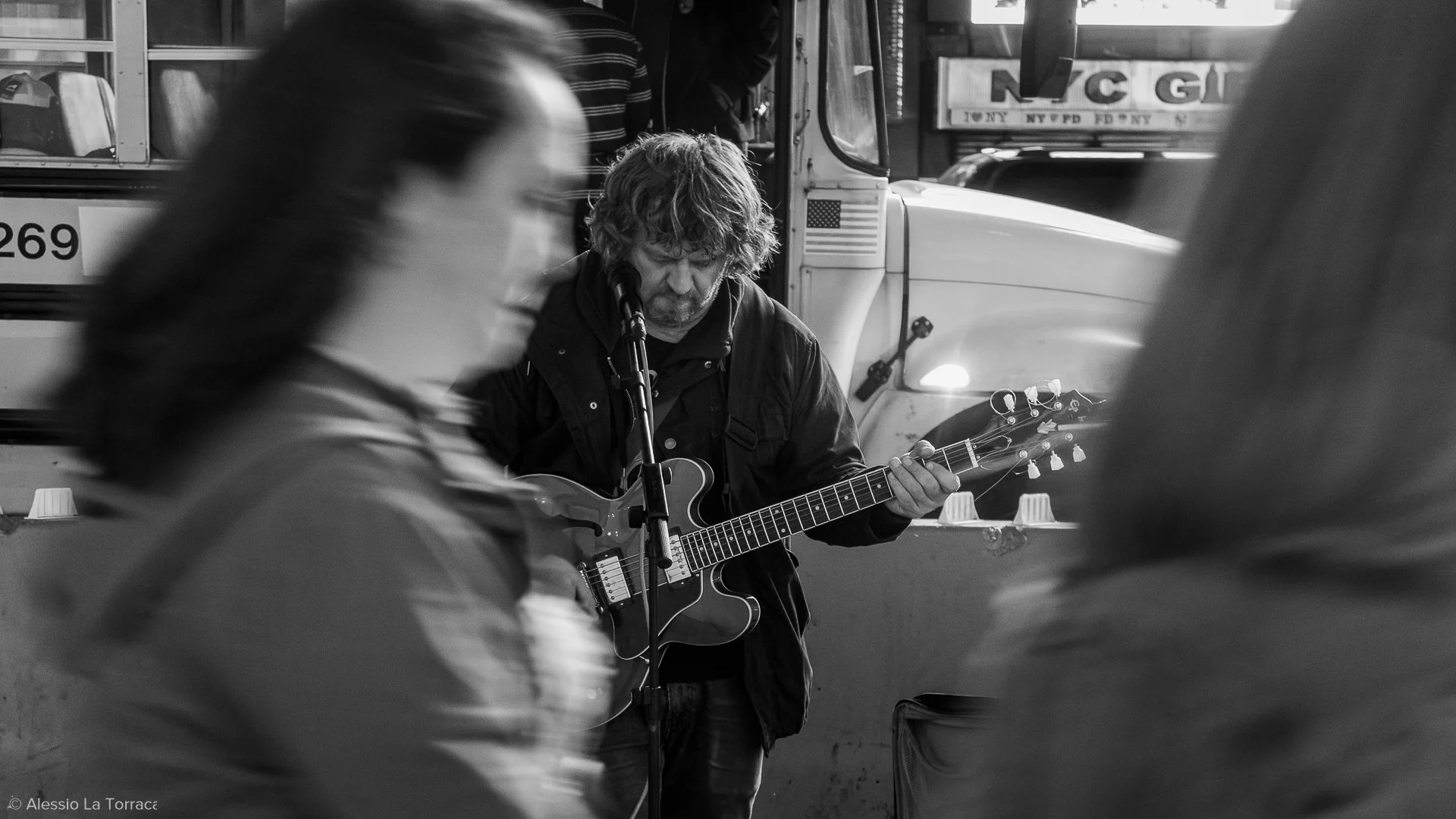 A street musician playing an electric guitar and singing into a microphone while walking past blurred pedestrians in an urban setting.