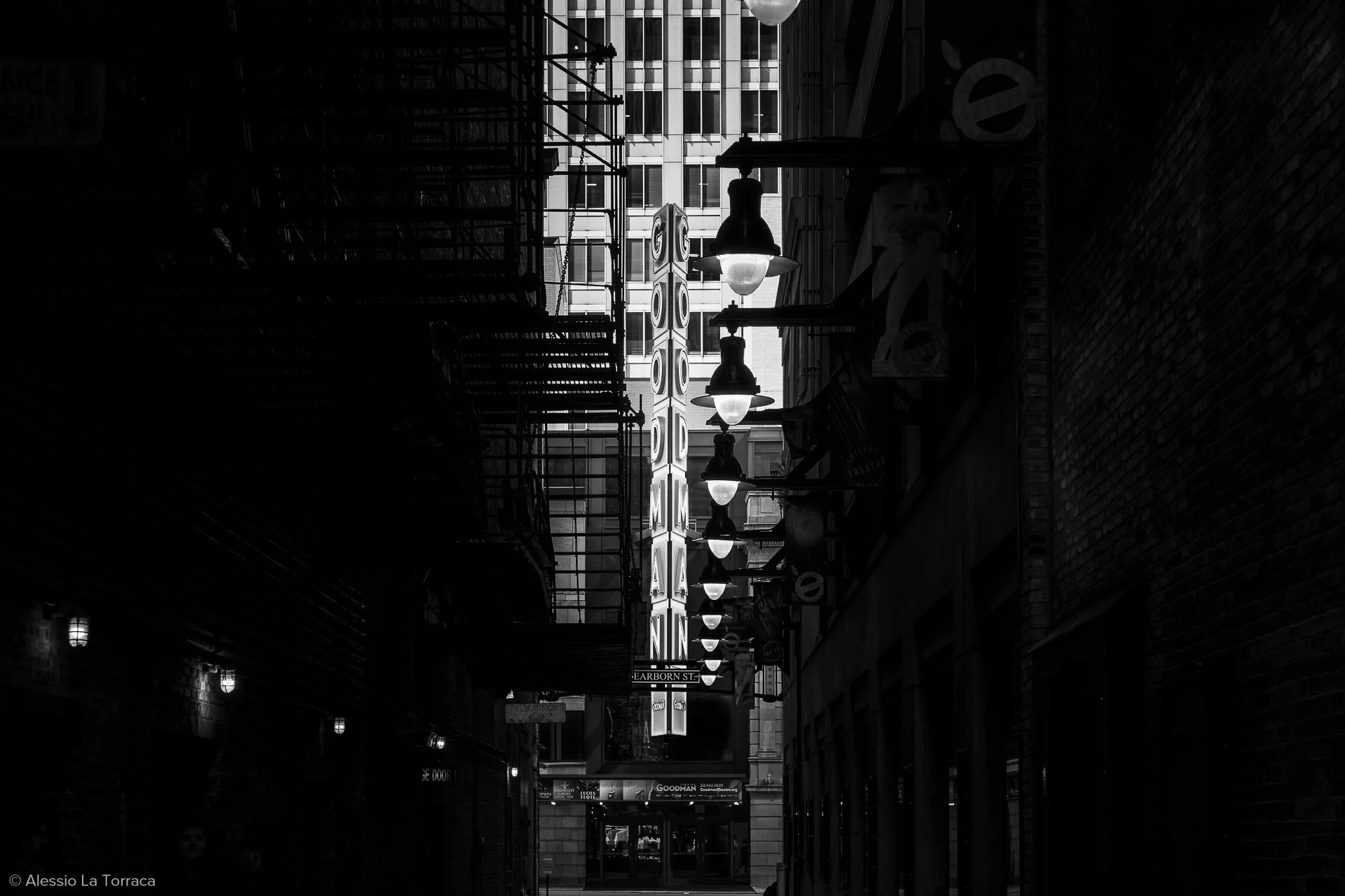 Black and white photo of a narrow city alley with illuminated signs, including a vertical sign reading "GOD ZILLA" and the Cimmaron Theatre sign, flanked by brick and building facades, with streetlights hanging overhead.