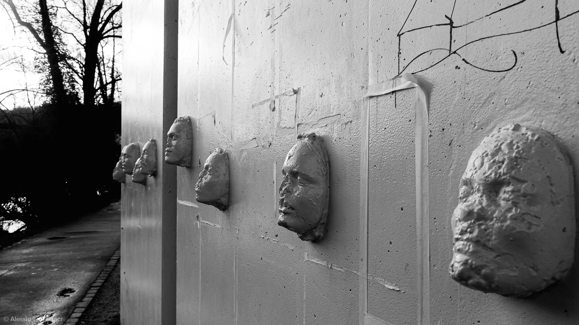 Black and white photo of a wall with multiple sculpted gray face busts attached to it, aligned in a row, with trees and a sidewalk in the background.