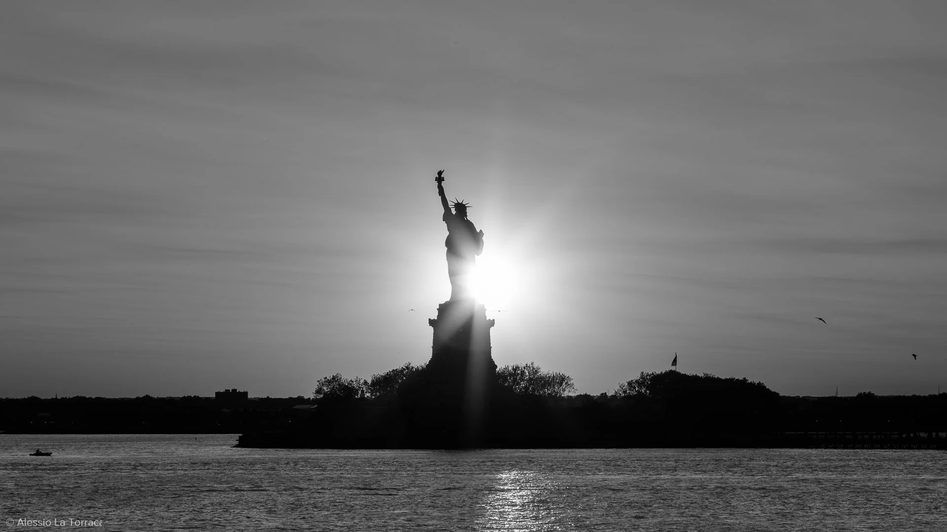 Silhouette of the Statue of Liberty at sunset with the sun behind it, reflecting on the water in the foreground.