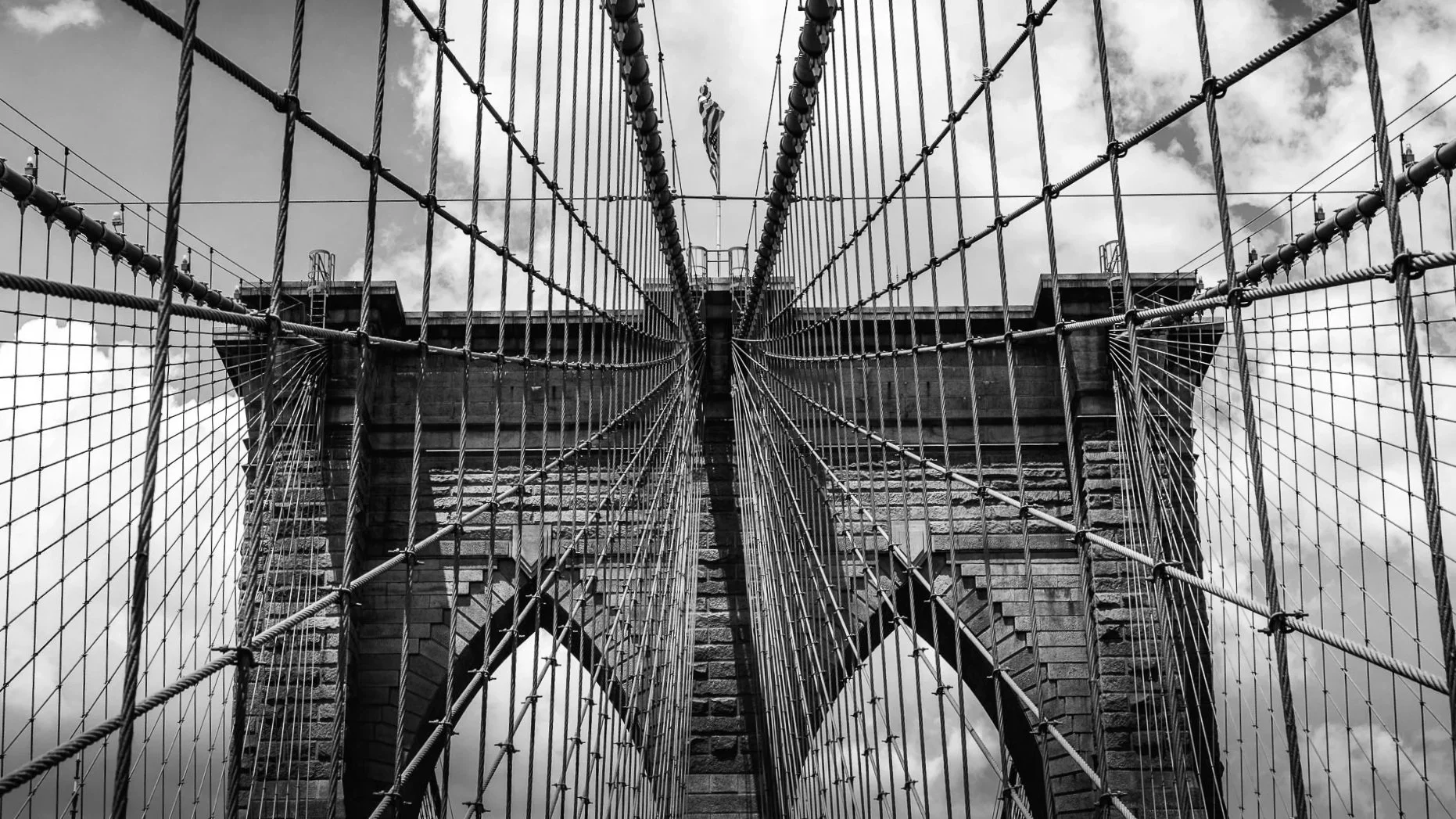 Looking up at the Brooklyn Bridge in New York City from the pedestrian walkway, showing the intricate steel cables and stone towers with an American flag hanging in the center, under a cloudy sky.