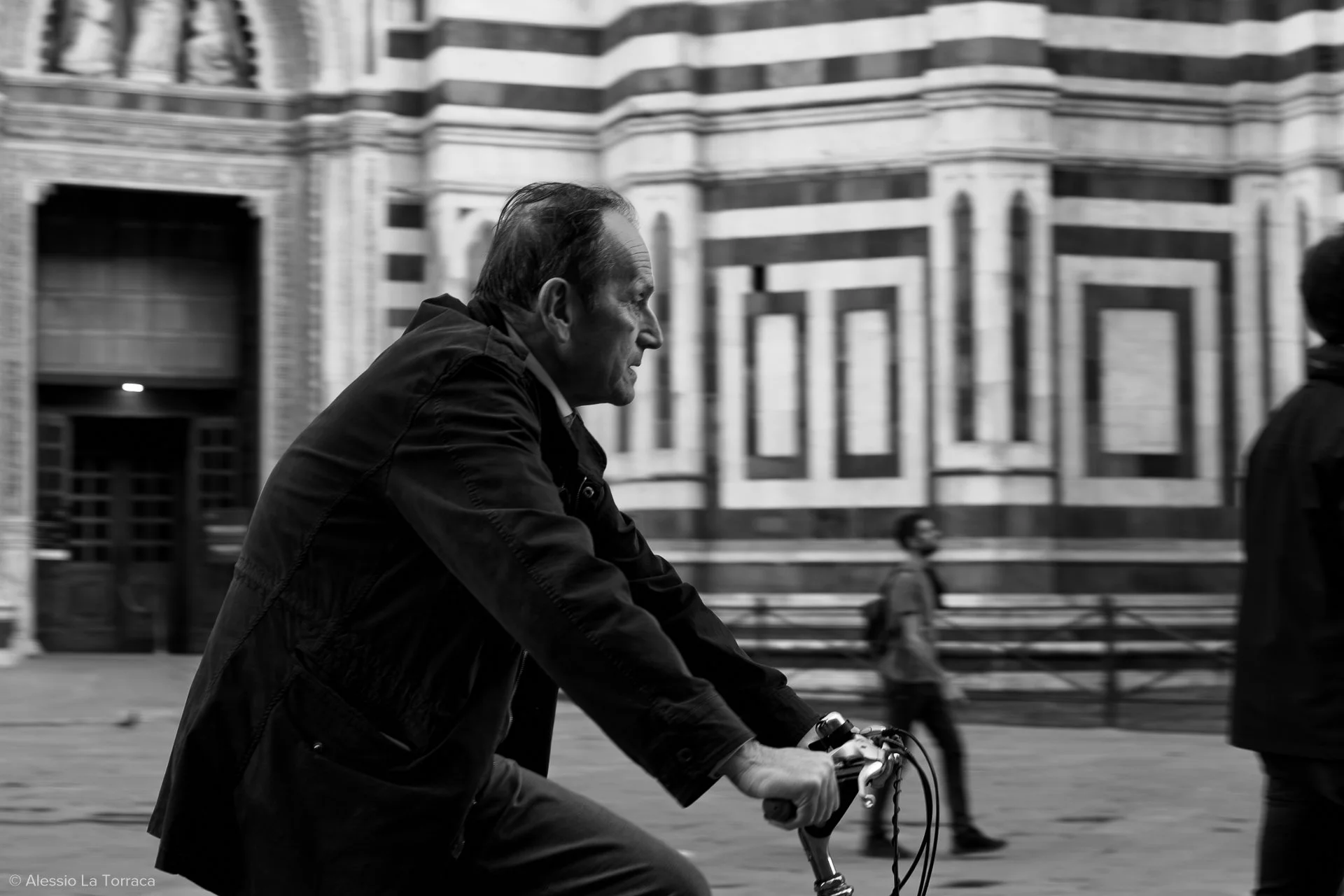 A black and white photo of a man riding a bicycle in front of a detailed, ornate building. The man has a serious expression and is wearing a dark jacket. There are blurred pedestrians in the background.