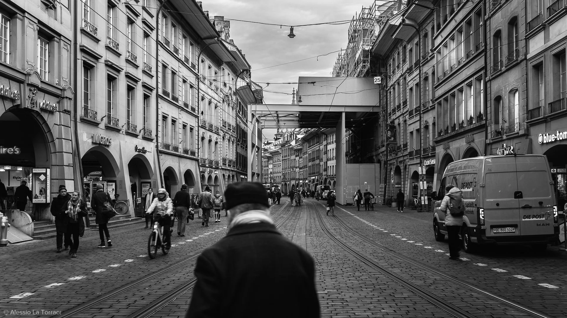 A bustling city street in black and white with pedestrians walking along cobblestone pavement and tram tracks. Classic European-style buildings line the street, with some under construction or renovation. A man in a coat and hat is in the foreground, walking away from the camera. Vehicles, including a postal truck, are parked along the street, and there are overhead wires and streetlights.